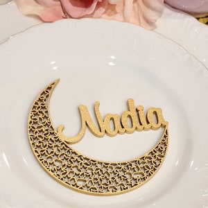 May include: A gold wooden crescent moon with a star pattern and the name Nadia written in cursive on top. The moon is on a white plate.