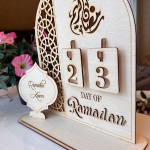 May include: A wooden Ramadan calendar with a decorative arch design. The calendar features a flip-style design with the numbers 2 and 3 displayed. The text "DAY OF RAMADAN" is written below the numbers. A separate wooden sign reads "Ramadan Kareem".