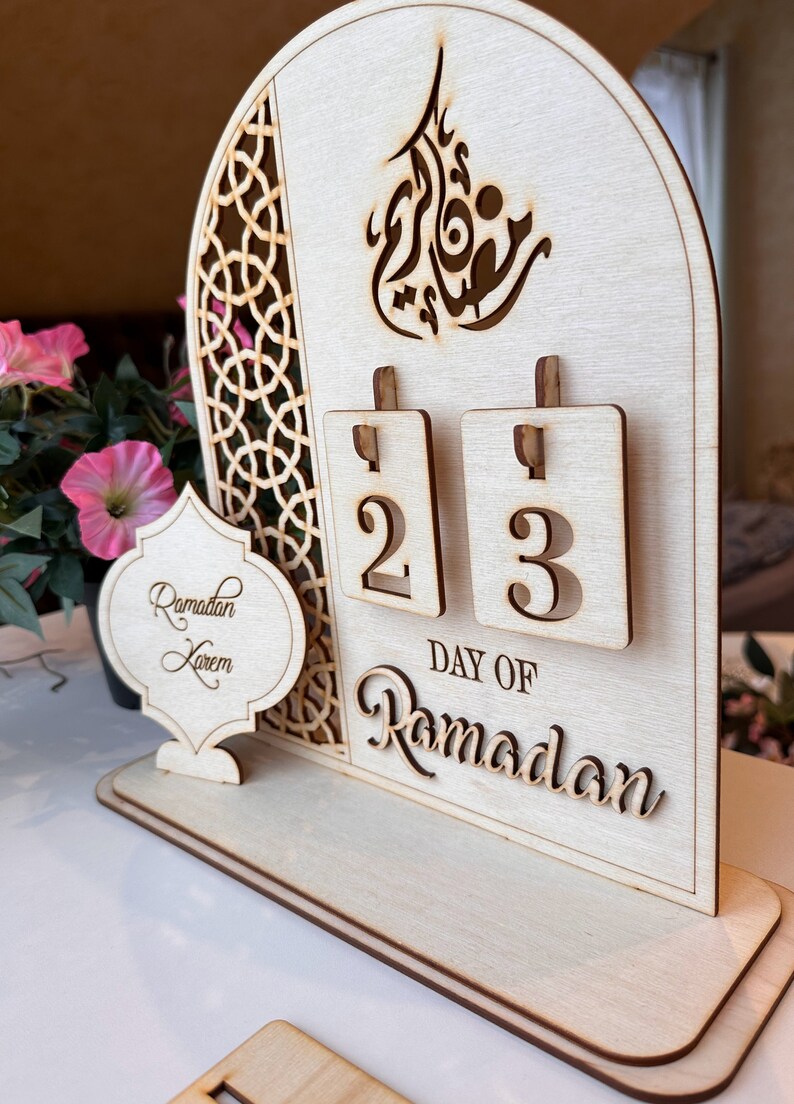 May include: A wooden Ramadan calendar with a decorative arch design. The calendar features a flip-style design with the numbers 2 and 3 displayed. The text "DAY OF RAMADAN" is written below the numbers. A separate wooden sign reads "Ramadan Kareem".