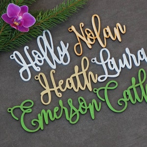May include: Five wooden names, "Holly", "Nolan", "Heath", "Laura", and "Emerson", are cut out in a cursive font. The names are in different colors: white, orange, gold, silver, and green.