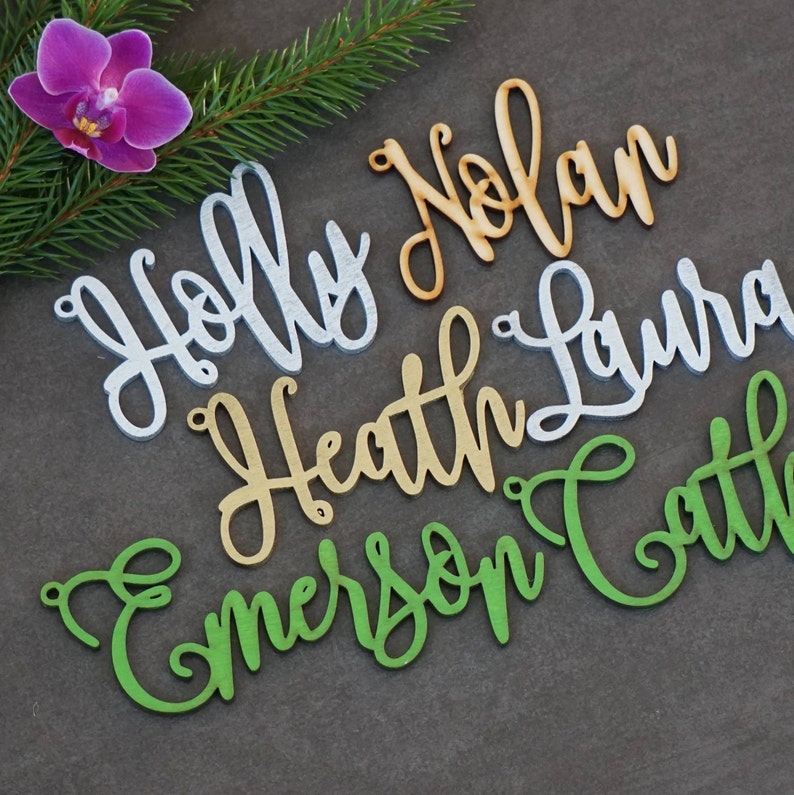 May include: Five wooden names, "Holly", "Nolan", "Heath", "Laura", and "Emerson", are cut out in a cursive font. The names are in different colors: white, orange, gold, silver, and green.