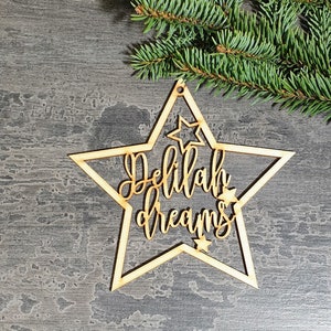 Puede incluir: Un adorno de madera en forma de estrella con las palabras "Delilah dreams" recortadas en una fuente cursiva. El adorno tiene un peque&ntilde;o agujero en la parte superior para colgar y est&aacute; rodeado de peque&ntilde;os recortes de estrellas. Una ramita de hoja perenne est&aacute; en el fondo.