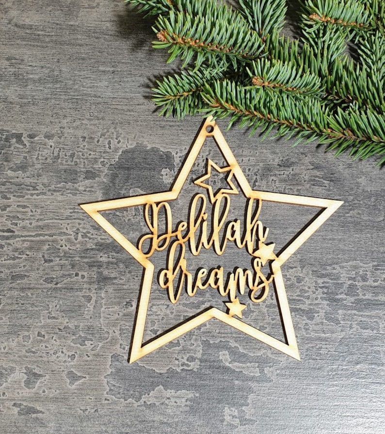 Puede incluir: Un adorno de madera en forma de estrella con las palabras "Delilah dreams" recortadas en una fuente cursiva. El adorno tiene un peque&ntilde;o agujero en la parte superior para colgar y est&aacute; rodeado de peque&ntilde;os recortes de estrellas. Una ramita de hoja perenne est&aacute; en el fondo.