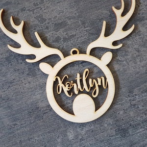 May include: A wooden Christmas ornament in the shape of a reindeer head with antlers. The name "Kortlyn" is cut out in a cursive font within the reindeer's face. The ornament is made of light-colored wood and has a small hole for hanging.