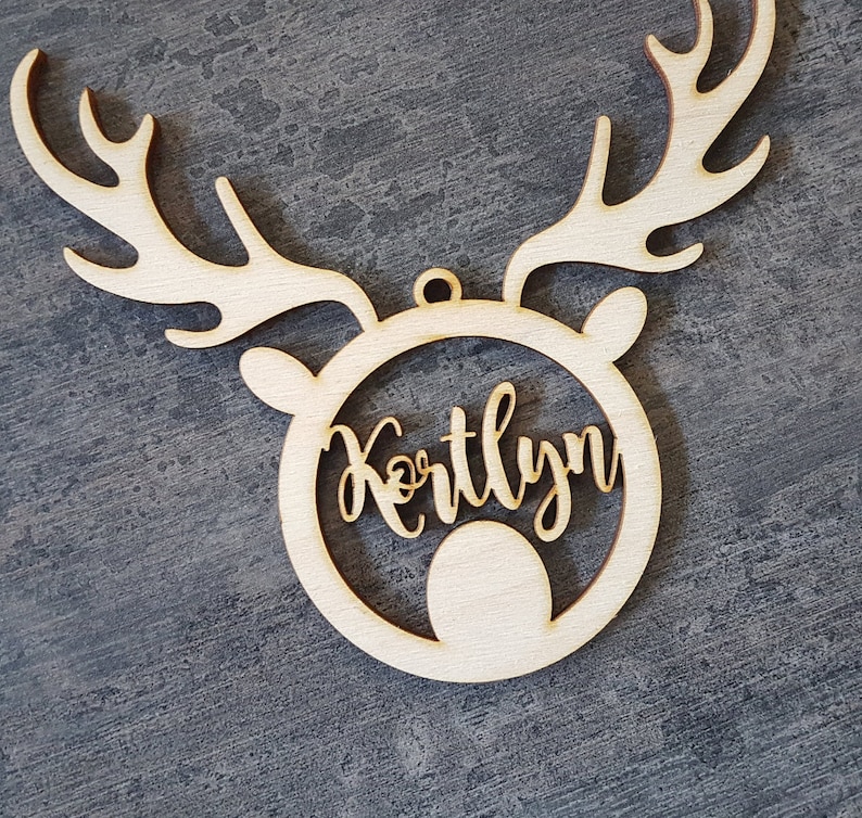 May include: A wooden Christmas ornament in the shape of a reindeer head with antlers. The name "Kortlyn" is cut out in a cursive font within the reindeer's face. The ornament is made of light-colored wood and has a small hole for hanging.