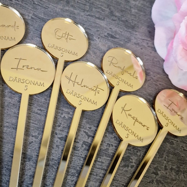 May include: Gold-colored acrylic drink stirrers with round, mirrored tops. Each top has a name written in cursive script and the text "DARSONAM 5". The names are: Otto, Irena, Rafaels, Helmuts, Ivars, and Kaspars.