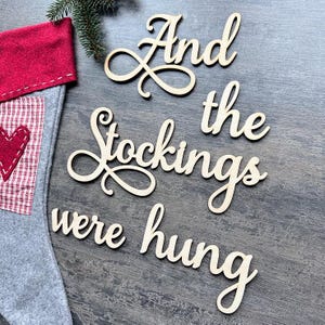 Può includere: Una calza di Natale grigia con polsino rosso e applicazione a cuore rosso, accanto a ritagli di legno che recitano "And the Stockings were hung". È presente anche un rametto di sempreverde.
