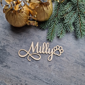 May include: Wooden sign with the name "Milly" and a paw print, on a gray background with gold wrapped ornaments and a pine branch.