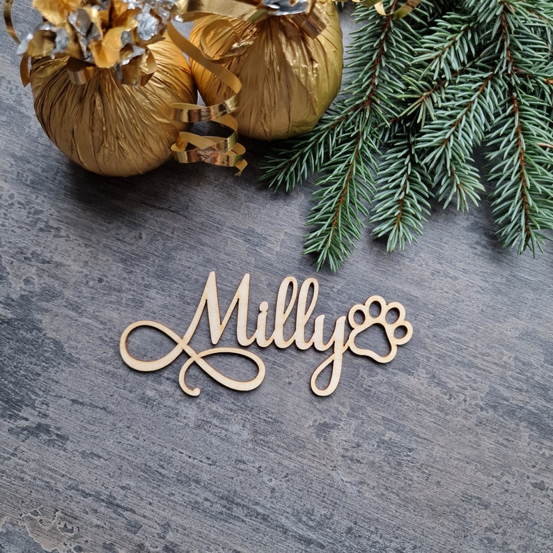 May include: Wooden sign with the name "Milly" and a paw print, on a gray background with gold wrapped ornaments and a pine branch.