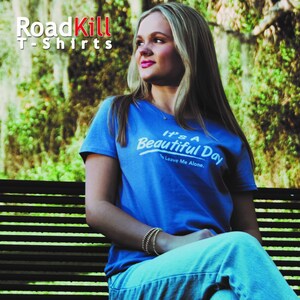 May include: A woman wearing a blue t-shirt with the text "It's a Beautiful Day to Leave Me Alone." The t-shirt is part of a collection called "RoadKill T-Shirts".