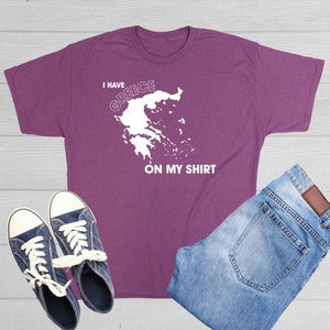 May include: A purple t-shirt with the text "I HAVE GREECE ON MY SHIRT" and a white outline of the country of Greece. The shirt is paired with blue jeans and navy blue sneakers with white laces.