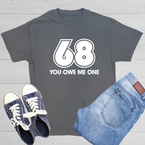 May include: A dark gray t-shirt with white text and a large white number 68. The text reads "You owe me one".