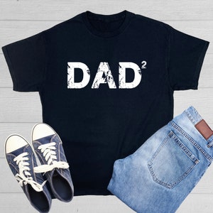May include: Black t-shirt with white distressed text that reads "DAD²". The shirt is laid flat on a woodgrain surface with a pair of blue sneakers and blue jeans.