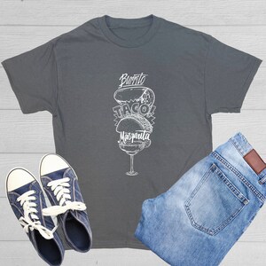May include: A charcoal gray t-shirt with a white graphic of a burrito, taco, and margarita glass. The shirt is paired with blue denim jeans and navy blue canvas sneakers with white laces. The items are arranged on a light wood surface.