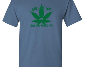 May include: A slate blue short-sleeve t-shirt featuring a green cannabis leaf graphic in the center. The text "ROLL ME UP" is printed above the leaf, "AND" is in the middle, and "SMOKE ME WHEN I DIE" is printed below the leaf in green.