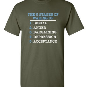 May include: A green t-shirt with white text that reads "THE 5 STAGES OF WAKING UP" and lists the stages as: 1. Denial, 2. Anger, 3. Bargaining, 4. Depression, 5. Acceptance.