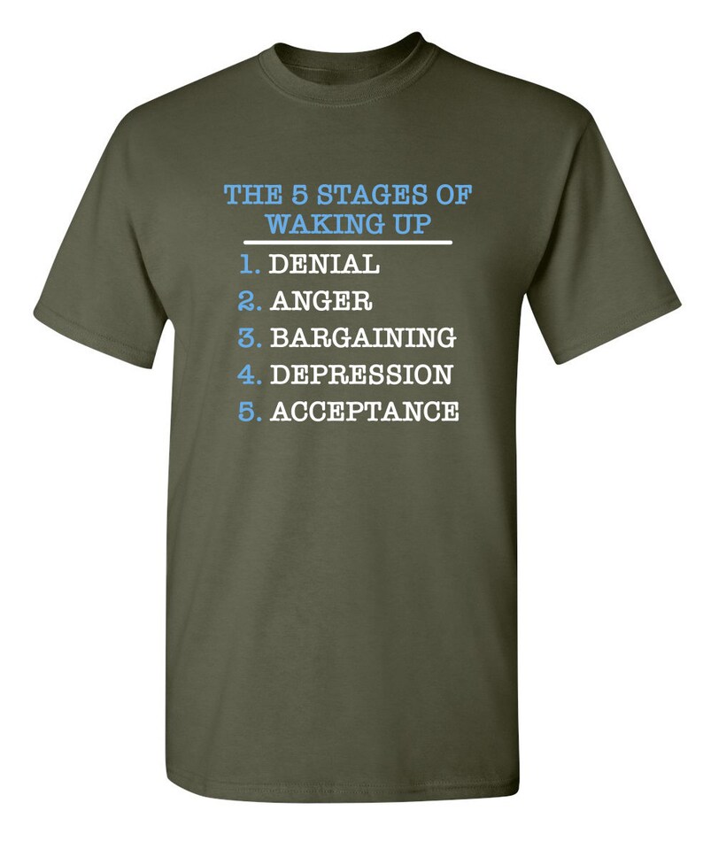 May include: A green t-shirt with white text that reads "THE 5 STAGES OF WAKING UP" and lists the stages as: 1. Denial, 2. Anger, 3. Bargaining, 4. Depression, 5. Acceptance.