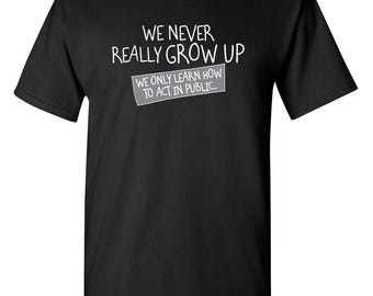 May include: A black short-sleeved t-shirt featuring white text that reads WE NEVER REALLY GROW UP and a secondary white text box that reads WE ONLY LEARN HOW TO ACT IN PUBLIC. The shirt has a crew neckline and a standard fit.