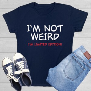 May include: A navy blue t-shirt with white text that reads "I'm not weird I'm limited edition!" The shirt is shown on a white wood background with a pair of blue jeans and blue sneakers.