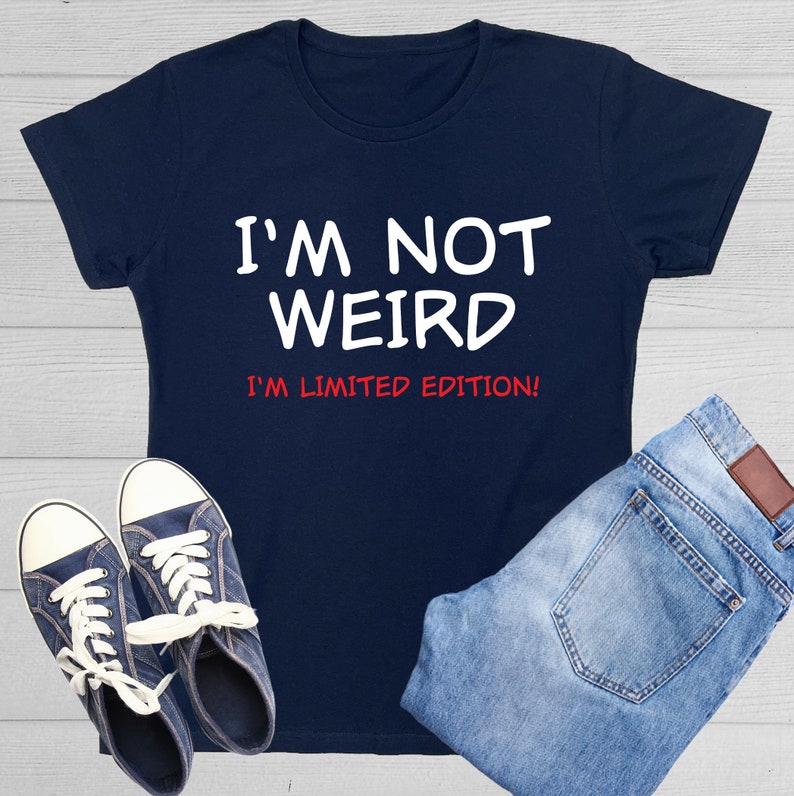 May include: A navy blue t-shirt with white text that reads "I'm not weird I'm limited edition!" The shirt is shown on a white wood background with a pair of blue jeans and blue sneakers.