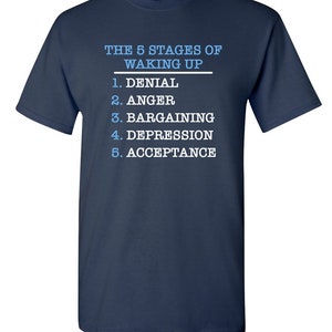 May include: A navy blue t-shirt with white text that reads "THE 5 STAGES OF WAKING UP" and lists the stages as: 1. Denial, 2. Anger, 3. Bargaining, 4. Depression, 5. Acceptance.