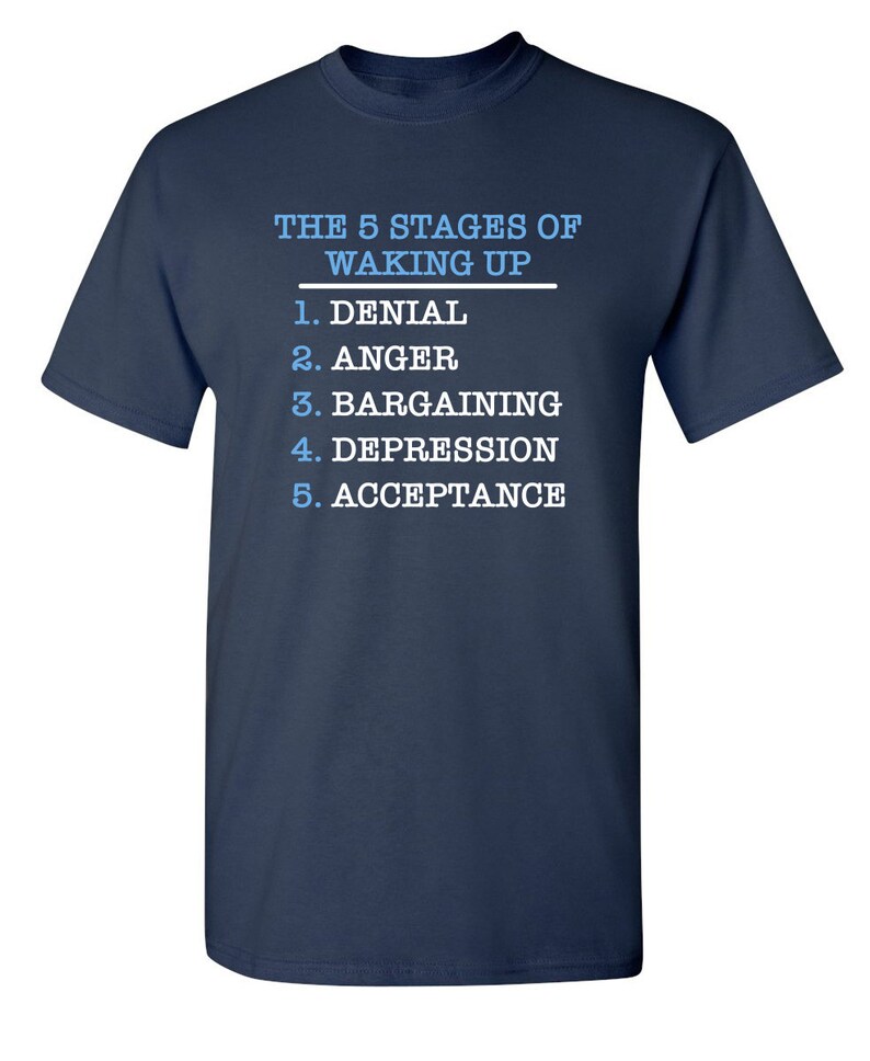 May include: A navy blue t-shirt with white text that reads "THE 5 STAGES OF WAKING UP" and lists the stages as: 1. Denial, 2. Anger, 3. Bargaining, 4. Depression, 5. Acceptance.
