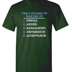 May include: A dark green t-shirt with white text that reads "THE 5 STAGES OF WAKING UP" and a list of the five stages: Denial, Anger, Bargaining, Depression, Acceptance.