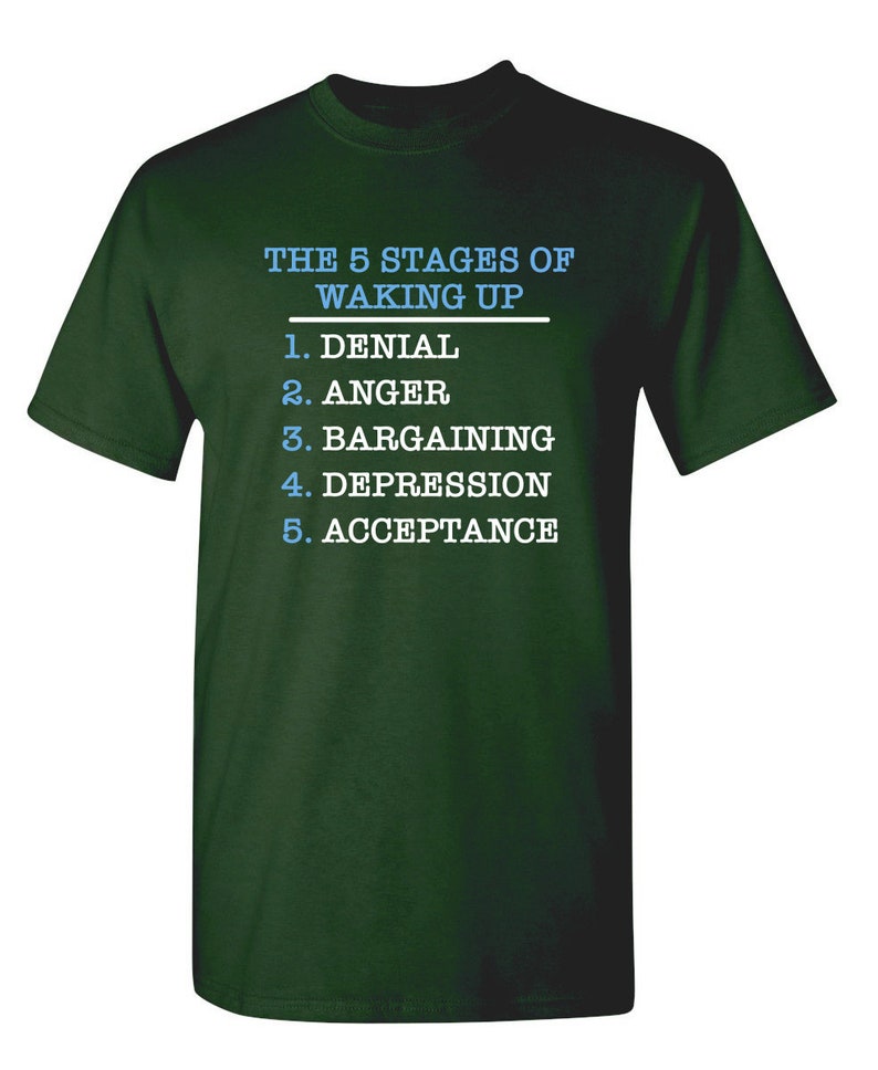 May include: A dark green t-shirt with white text that reads "THE 5 STAGES OF WAKING UP" and a list of the five stages: Denial, Anger, Bargaining, Depression, Acceptance.