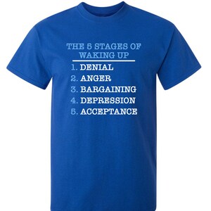 May include: A royal blue t-shirt with white text that reads "THE 5 STAGES OF WAKING UP" and lists the stages as: 1. Denial, 2. Anger, 3. Bargaining, 4. Depression, 5. Acceptance.