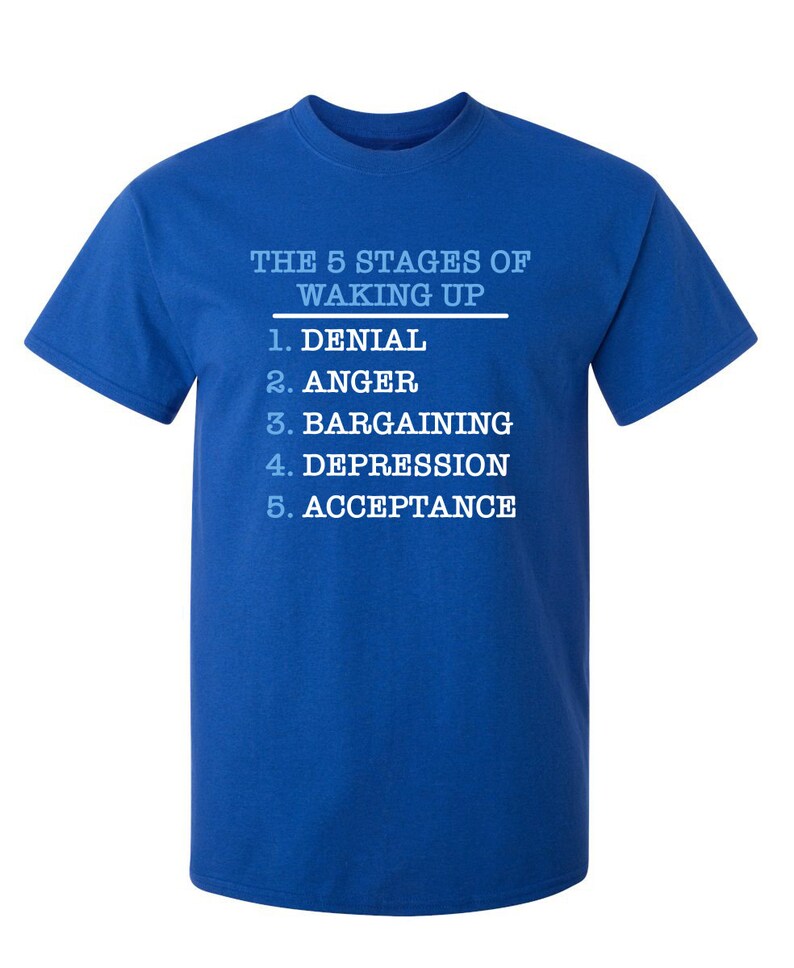 May include: A royal blue t-shirt with white text that reads "THE 5 STAGES OF WAKING UP" and lists the stages as: 1. Denial, 2. Anger, 3. Bargaining, 4. Depression, 5. Acceptance.
