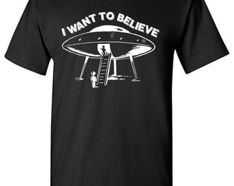 May include: A black short-sleeved t-shirt featuring a white graphic of a flying saucer with a ladder extended to the ground and a figure standing nearby. The text I WANT TO BELIEVE is printed in white in an arc above the flying saucer illustration.