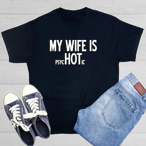 May include: Black t-shirt with the white text "MY WIFE IS PSYCHOTIC HOT" displayed prominently. The shirt is paired with blue jeans and navy blue sneakers with white laces, creating a casual ensemble.