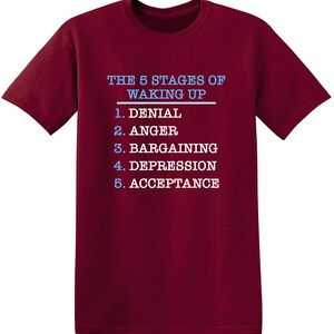 May include: Burgundy t-shirt with white text that reads "THE 5 STAGES OF WAKING UP" and a list of the stages: 1. Denial, 2. Anger, 3. Bargaining, 4. Depression, 5. Acceptance.