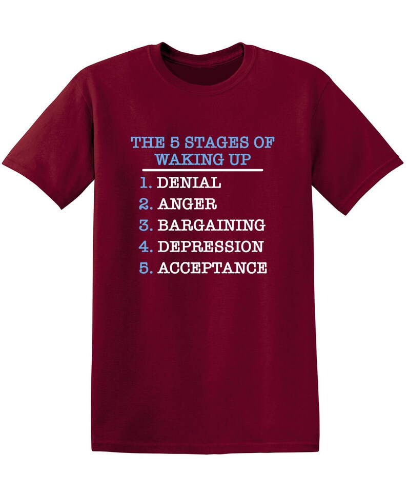 May include: Burgundy t-shirt with white text that reads "THE 5 STAGES OF WAKING UP" and a list of the stages: 1. Denial, 2. Anger, 3. Bargaining, 4. Depression, 5. Acceptance.
