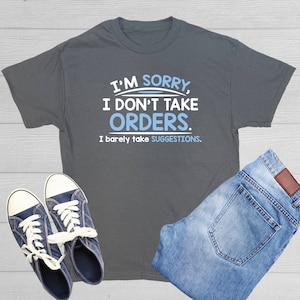 May include: A dark gray t-shirt with the text "I'm sorry, I don't take orders. I barely take suggestions." in white text with a blue underline under "orders".