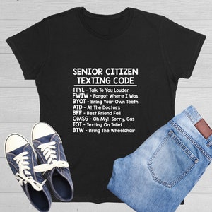 May include: Black t-shirt with the text "SENIOR CITIZEN TEXTING CODE" and a list of texting acronyms. The shirt is paired with blue jeans and navy blue sneakers with white laces.