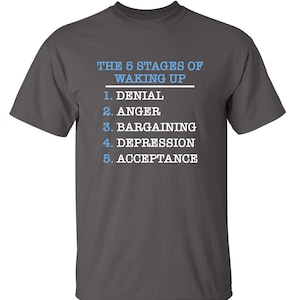 May include: A dark gray t-shirt with white text that reads "THE 5 STAGES OF WAKING UP" and a list of the five stages: Denial, Anger, Bargaining, Depression, and Acceptance.