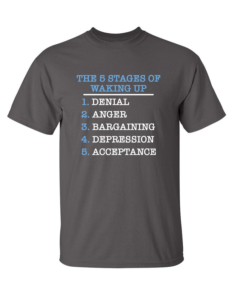 May include: A dark gray t-shirt with white text that reads "THE 5 STAGES OF WAKING UP" and a list of the five stages: Denial, Anger, Bargaining, Depression, and Acceptance.