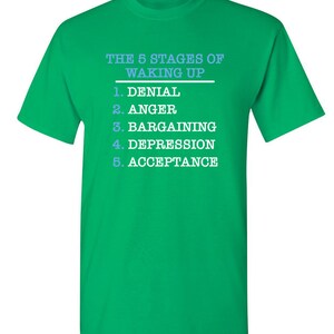 May include: Green t-shirt with white text that reads "The 5 Stages of Waking Up" and lists the stages as: 1. Denial, 2. Anger, 3. Bargaining, 4. Depression, 5. Acceptance.