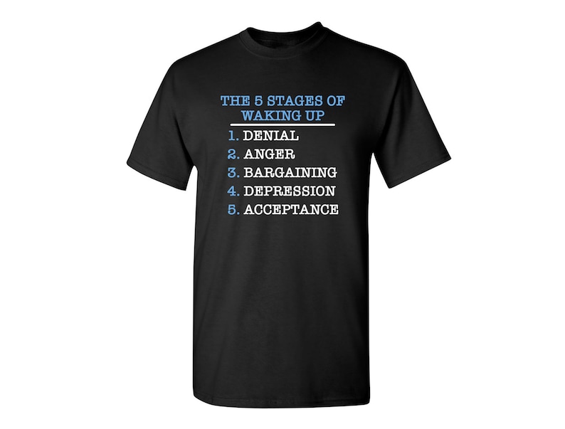 May include: Black t-shirt with white text that reads "THE 5 STAGES OF WAKING UP" and a list of the stages: Denial, Anger, Bargaining, Depression, Acceptance.