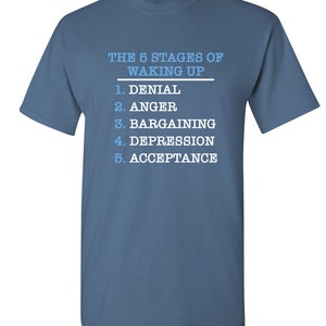 May include: A blue t-shirt with white text that reads "THE 5 STAGES OF WAKING UP" and lists the stages as: 1. Denial, 2. Anger, 3. Bargaining, 4. Depression, 5. Acceptance.