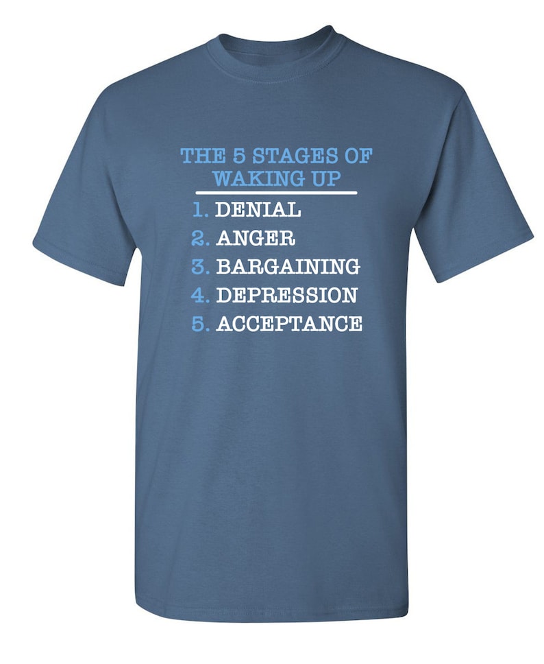 May include: A blue t-shirt with white text that reads "THE 5 STAGES OF WAKING UP" and lists the stages as: 1. Denial, 2. Anger, 3. Bargaining, 4. Depression, 5. Acceptance.