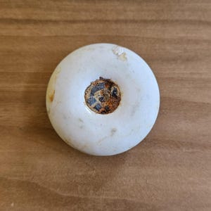 May include: A round, white ceramic knob with a smooth, slightly weathered surface. The center features a circular, recessed metal insert that shows signs of oxidation, rust, and dark brown discoloration, contrasting with the white finish.