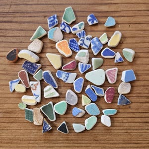 Puede incluir: Una colección de fragmentos de vidrio marino de diversas formas y tamaños, dispersos sobre una superficie de madera. Las piezas muestran una gama de colores, incluyendo azul, verde, blanco, amarillo y rojo, con algunos diseños intrincados. El vidrio marino parece provenir de cerámica rota.