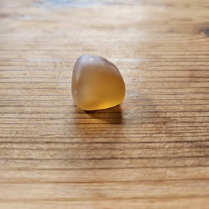 May include: A polished, amber-toned gemstone with a smooth, rounded shape. The translucent stone sits on a wooden surface, displaying visible wood grain. The stone's colour is a warm, golden hue.