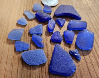 May include: A collection of sea glass pieces in various shades of blue, ranging from light to deep indigo, scattered on a wooden surface. A silver coin is also present. The sea glass pieces vary in shape and size, with smooth, frosted textures.