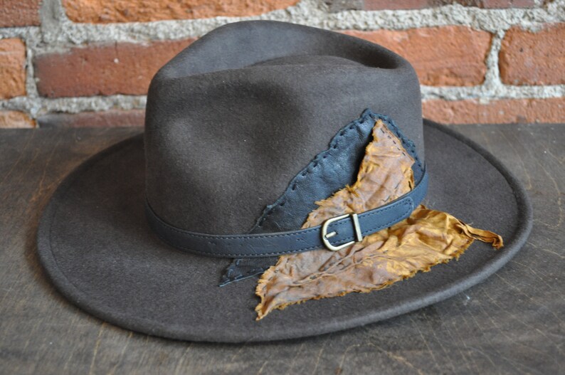 May include: A brown felt fedora hat with a black leather band and a brown leather leaf accent.