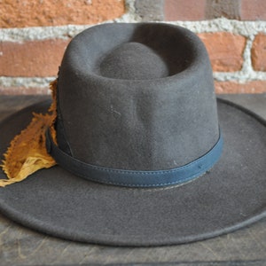 May include: A dark gray fedora hat with a wide brim and a blue leather band. The hat has a decorative piece of brown leather attached to the side.