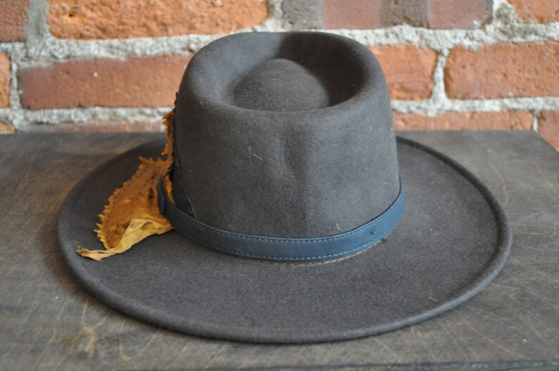 May include: A dark gray fedora hat with a wide brim and a blue leather band. The hat has a decorative piece of brown leather attached to the side.