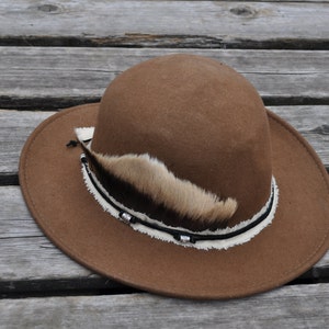 Puede incluir: Un sombrero fedora de fieltro marrón con una ala ancha y una banda decorativa de tela blanca y cuero negro. La banda está adornada con una pequeña pluma y cuentas.